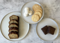Three plates with sliced brown bread, cookies, and cream on a textured surface