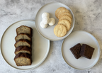 Three plates with sliced brown bread, cookies, and cream on a textured surface