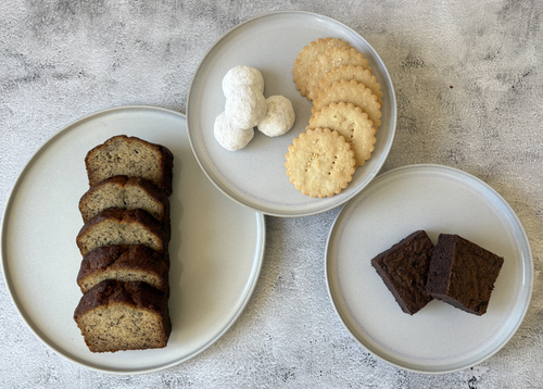 Three plates with sliced brown bread, cookies, and cream on a textured surface