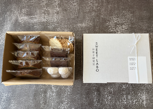 Box of assorted pastries next to a white box with 'SWEET LABO' branding on a textured gray surface.